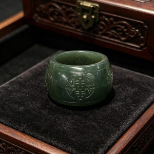 May include: A dark green jade ring with carved symbols. The ring is cylindrical with a flat top and bottom. The carved symbols are visible around the ring's exterior. The ring is displayed on a black velvet surface inside a wooden box.