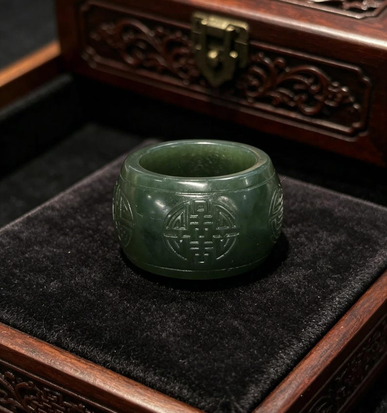May include: A dark green jade ring with carved symbols. The ring is cylindrical with a flat top and bottom. The carved symbols are visible around the ring's exterior. The ring is displayed on a black velvet surface inside a wooden box.