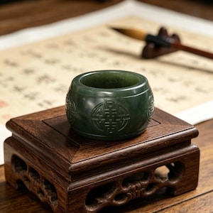 May include: A dark green jade ring with carved symbols sits on a small, dark wooden stand. The ring is cylindrical with a smooth, polished surface. The stand has a square top and ornate, carved sides. A calligraphy brush and paper are in the background.