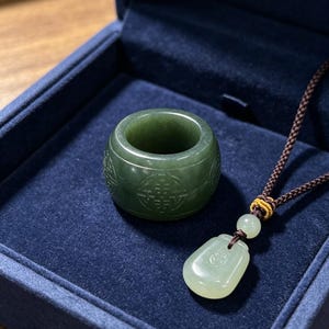 May include: A jade ring and pendant necklace set. The ring is a dark green colour with carved patterns. The pendant is a lighter green, teardrop-shaped, and hangs from a brown cord. The set is displayed in a dark blue box.