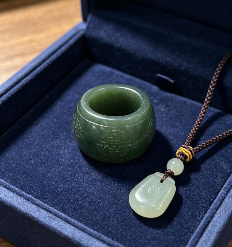 May include: A jade ring and pendant necklace set. The ring is a dark green colour with carved patterns. The pendant is a lighter green, teardrop-shaped, and hangs from a brown cord. The set is displayed in a dark blue box.