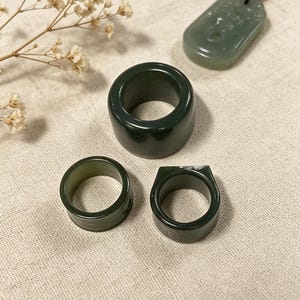 以下が含まれることがあります： さまざまなデザインの濃い緑色の翡翠の指輪が3つ表示されています。1つは太いバンド、もう1つは細いバンド、3つ目は長方形のトップです。彫刻が施されたデザインの翡翠のペンダントも存在します。