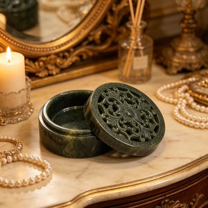 K&ouml;nnte beinhalten: Eine offene, dunkelgr&uuml;ne Jade-Box mit einem Deckel mit filigranem floralem Design. Die Box steht auf einer Marmoroberfl&auml;che, mit einer brennenden Kerze und Schmuck im Hintergrund. Die Box ist ein dekorativer Gegenstand.