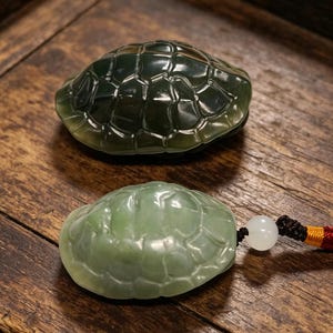 May include: Two jade turtle figurines. The top turtle is dark green, and the bottom turtle is light green. The bottom turtle has a white bead and a red, yellow, and black cord attached. Both turtles are on a wooden surface.
