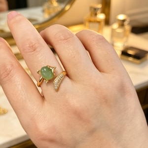 Op de afbeelding: Een goudkleurige ring met een lichtgroene ovale edelsteen en een halvemaanvormige band versierd met kleine, heldere kristallen. De ring zit om een vinger, met een vage achtergrond.