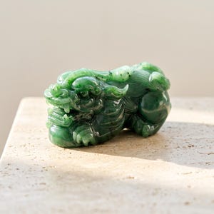 Puede incluir: Una figura de jade verde tallada que representa a una criatura mítica similar a un león en posición agachada. La escultura presenta detalles intrincados en la cara, la melena y el cuerpo, con una superficie lisa y pulida que muestra variaciones.