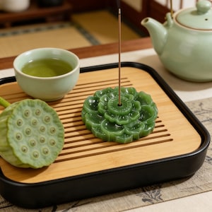 May include: A green lotus-shaped incense holder with an incense stick, a cup of green tea, and a lotus seed pod on a bamboo tray. A green teapot is in the background. The tray has a black border.