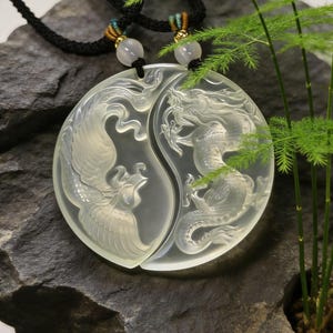 May include: A clear, carved pendant necklace featuring a phoenix and dragon design, symbolizing balance. The pendant is split into two halves, each depicting one of the mythical creatures. The necklace has a black cord with decorative beads and gold accents.