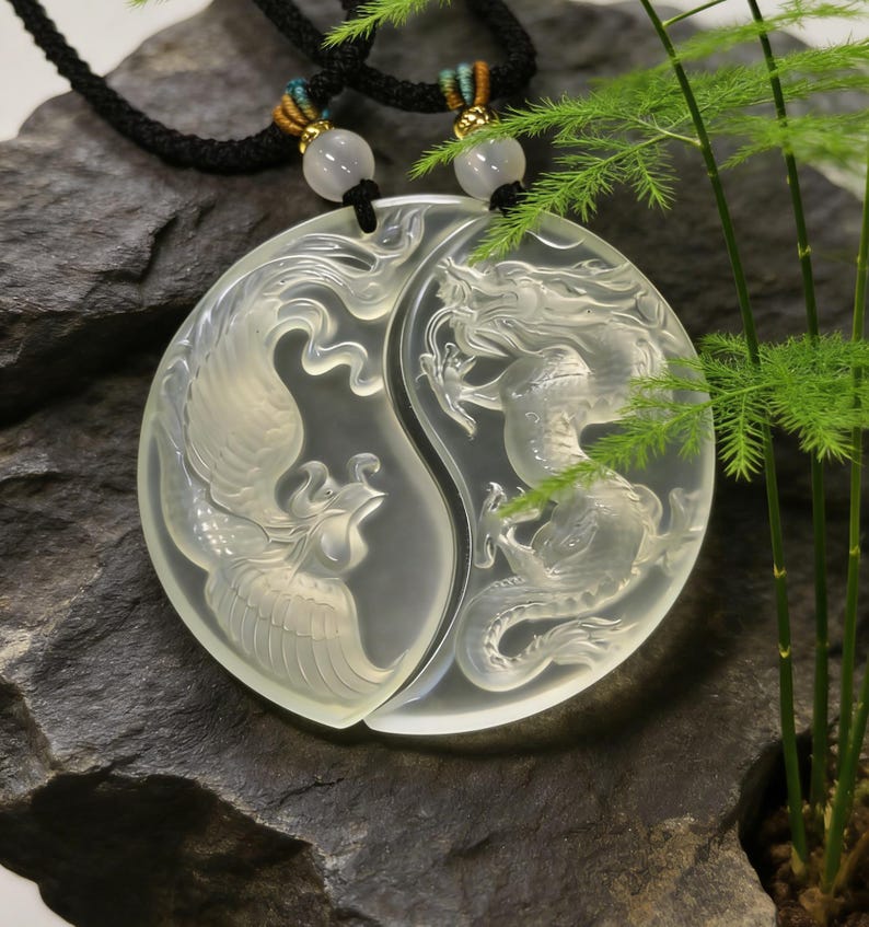 May include: A clear, carved pendant necklace featuring a phoenix and dragon design, symbolizing balance. The pendant is split into two halves, each depicting one of the mythical creatures. The necklace has a black cord with decorative beads and gold accents.