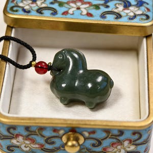 Op de afbeelding: Donkergroene jade hanger in de vorm van een paard, hangend aan een zwart koord met een rode kraal. De hanger ligt in een decoratieve doos met een blauw bloemmotief en gouden rand.