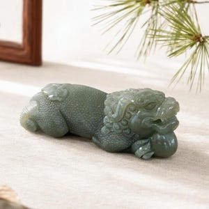 Può includere: Una statuetta in pietra scolpita color verde salvia che raffigura una creatura mitica in posizione sdraiata. La scultura presenta dettagli intricati sulla testa, sulla criniera e sul corpo testurizzato, con la creatura che poggia le zampe anteriori su un piccolo globo.