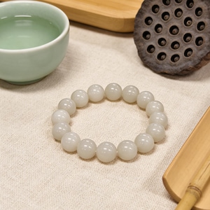 May include: A white beaded bracelet with large, round beads. The bracelet is resting on a cream-colored cloth, next to a light green teacup, a wooden tray, and a dried lotus seed pod. The bracelet is a simple, elegant piece of jewelry.