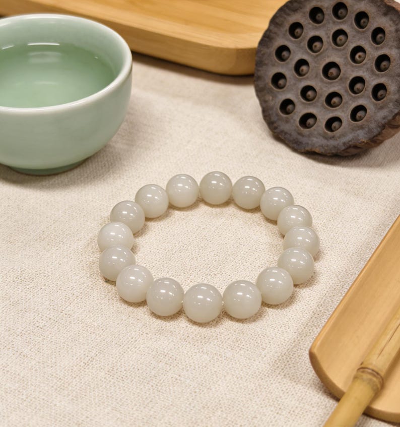 May include: A white beaded bracelet with large, round beads. The bracelet is resting on a cream-colored cloth, next to a light green teacup, a wooden tray, and a dried lotus seed pod. The bracelet is a simple, elegant piece of jewelry.