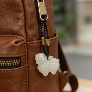 May include: A white jade horse pendant hangs from a black cord with gold accents, attached to a brown leather backpack. The horse figurine is detailed and smooth, with a polished finish. The backpack has a zipper and a small pocket.