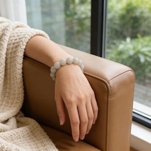 May include: A light green beaded bracelet with large, round beads. The bracelet is worn on a person's wrist, resting on a brown leather armchair. A cream-colored knitted blanket is draped over the arm of the chair.