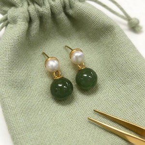 Può includere: Orecchini dorati con una perla bianca e una pietra rotonda verde scuro. Gli orecchini sono esposti su un sacchetto verde chiaro, con pinzette dorate in primo piano.