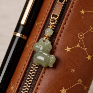 May include: A close-up of a brown leather pencil case with a gold zipper and a small, green jade horse charm. A black pen with gold accents is visible. The case has a constellation pattern in gold.