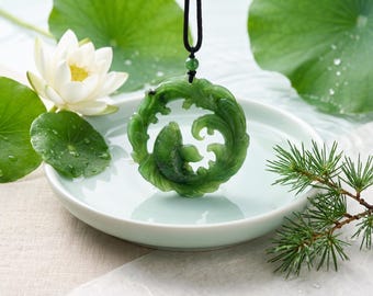 May include: A circular green jade pendant featuring an intricate carved design of a fish swimming amidst stylised waves. The pendant is suspended from a simple black cord and includes a small matching green jade bead above the main carving.