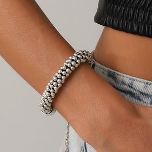 Może przedstawiać: Srebrna bransoletka z wzorem z koralików. Bransoletka wykonana jest z małych, okrągłych koralików i posiada zapięcie na łańcuszek. Bransoletka jest noszona na nadgarstku.