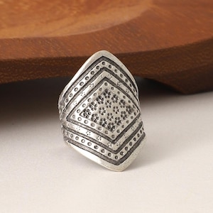 Peut inclure: Une bague argentée ornée d'un motif géométrique complexe. La bague présente un motif central en forme de losange entouré de rangées de petits points et de lignes. L'anneau est posé sur un fond en bois.