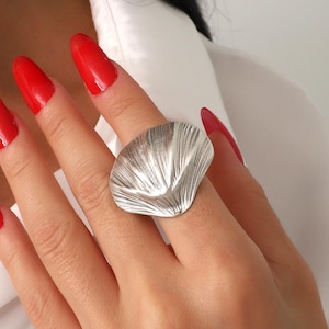 May include: A large, silver-colored ring with a textured, fan-like design. The ring is worn on a finger with red nail polish. The background is a white shirt.