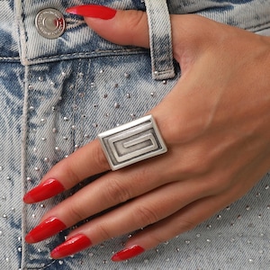 Puede incluir: Un anillo geométrico plateado con un diseño rectangular. El anillo presenta un patrón empotrado en forma de laberinto. La mano descansa sobre unos vaqueros de mezclilla descoloridos.