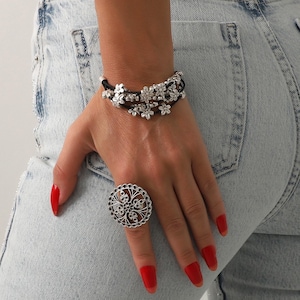 May include: A hand wearing a silver ring with an intricate floral design and a black cord bracelet adorned with silver flower charms. The person is wearing light blue jeans and has red nail polish.