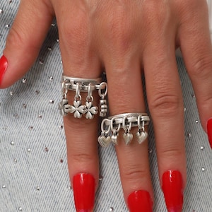 May include: Two silver rings with dangling charms, one with flowers and the other with hearts. The rings are worn on a hand with long, red fingernails. The background includes a light blue denim fabric.