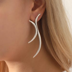 Peut inclure: Boucles d'oreilles argentées abstraites avec un design incurvé et fluide. Les boucles d'oreilles ont une esthétique moderne et minimaliste, avec une finition lisse et polie. Le design s'incurve vers le haut et vers l'extérieur, créant une forme unique et accrocheuse. Ces boucles d'oreilles sont une pièce de déclaration.