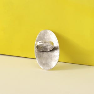 May include: A silver-toned ring with an oval face. The ring's face has a textured, hammered appearance. The band is a simple, thin design. The background is a bright yellow.