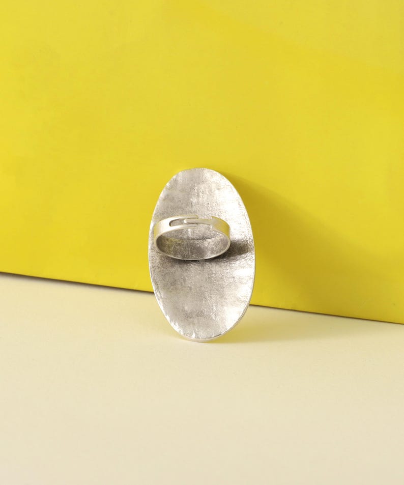 May include: A silver-toned ring with an oval face. The ring's face has a textured, hammered appearance. The band is a simple, thin design. The background is a bright yellow.
