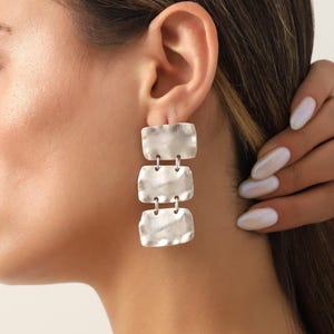May include: Silver-toned earrings with a textured, rectangular design. Each earring features three connected rectangular shapes, creating a dangling effect. The earrings are shown on a person's ear.