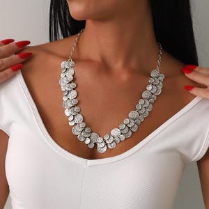May include: A silver-toned necklace featuring a cascade of textured, circular pendants. The necklace is worn against a white top, creating a contrast that highlights the jewelry's design. The chain is delicate, and the pendants vary in size.