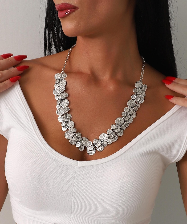 May include: A silver-toned necklace featuring a cascade of textured, circular pendants. The necklace is worn against a white top, creating a contrast that highlights the jewelry's design. The chain is delicate, and the pendants vary in size.