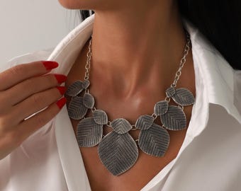 May include: Silver-toned statement necklace featuring leaf-shaped pendants with textured detailing. The necklace is worn over a white collared shirt. The model's red nail polish and lipstick add a pop of color.