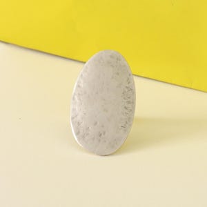 May include: A silver-colored, oval-shaped ring with a textured surface. The ring is set against a yellow and cream-colored background, creating a simple, yet elegant aesthetic. The ring's design is minimalist and modern.