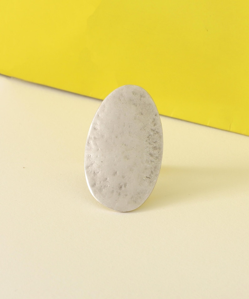 May include: A silver-colored, oval-shaped ring with a textured surface. The ring is set against a yellow and cream-colored background, creating a simple, yet elegant aesthetic. The ring's design is minimalist and modern.