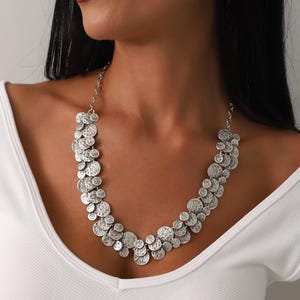 May include: Silver-toned statement necklace featuring a cluster of textured, circular pendants. The necklace is composed of multiple overlapping discs, creating a layered effect. The jewelry is worn against a white top.