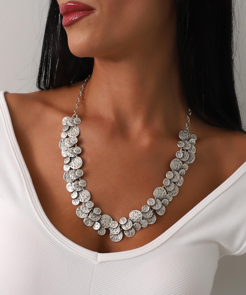 May include: Silver-toned statement necklace featuring a cluster of textured, circular pendants. The necklace is composed of multiple overlapping discs, creating a layered effect. The jewelry is worn against a white top.
