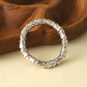 May include: A silver-colored bracelet with a textured, bamboo-like design. The bracelet is circular and appears to be made of individual, connected segments. The background includes a wooden bowl and a cream-colored surface.