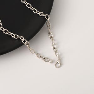 May include: Silver-toned chain necklace with a lobster clasp closure. The necklace features a series of oval-shaped links. The jewelry is displayed on a black plate against a white background.