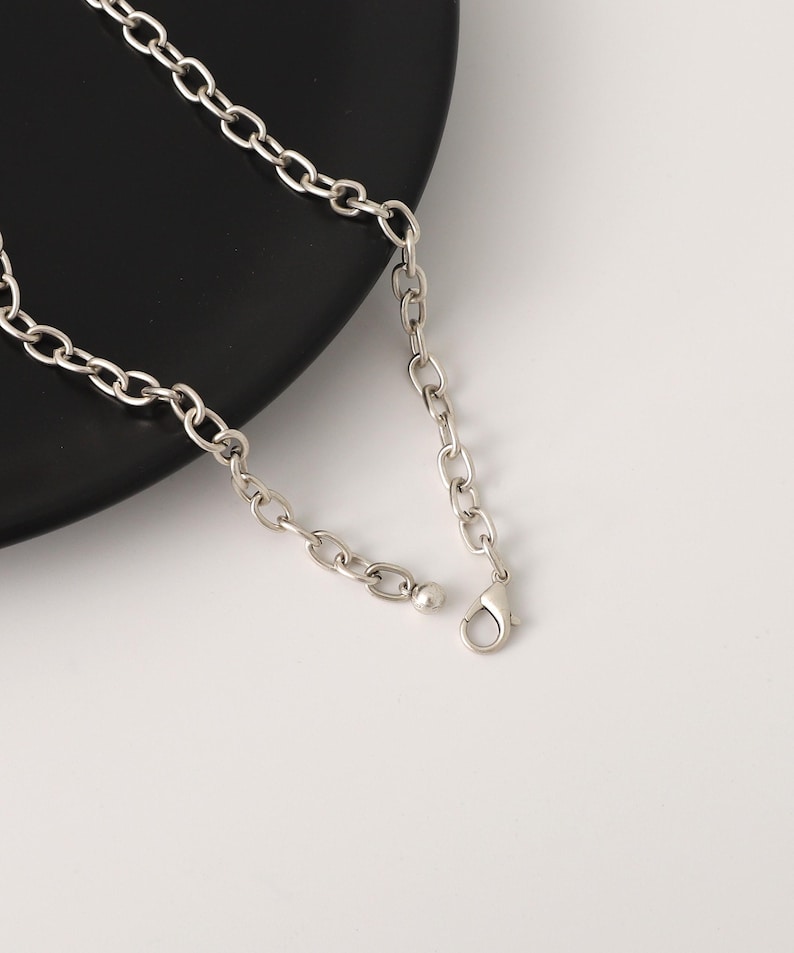 May include: Silver-toned chain necklace with a lobster clasp closure. The necklace features a series of oval-shaped links. The jewelry is displayed on a black plate against a white background.
