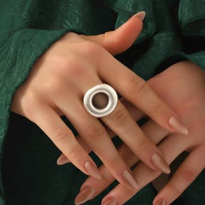 May include: A silver ring with a circular design. The ring has a solid outer band and a hollow centre. The ring is worn on a finger, with the hands and a green fabric background visible.
