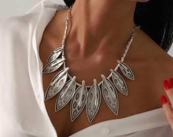 May include: A silver-toned statement necklace featuring a series of leaf-shaped pendants with a textured, oxidized finish. The necklace is composed of multiple pointed, petal-like elements arranged in a fan shape, connected by a simple silver chain.