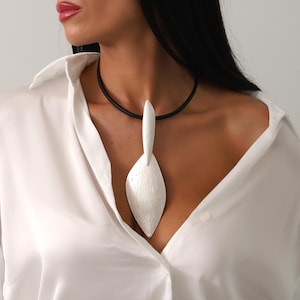 May include: A silver pendant necklace with a black cord. The pendant is leaf-shaped with a textured finish. The necklace is worn over a white button-down shirt.