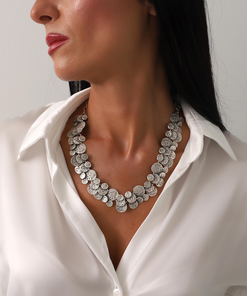 May include: Silver-toned statement necklace featuring a cluster of textured, circular pendants. The necklace is worn over a white button-down shirt. The pendants vary in size and have a hammered or embossed appearance, creating a unique, eye-catching design.