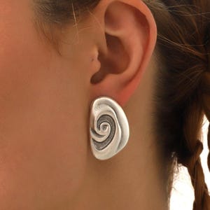 May include: A silver-toned, abstract stud earring with a spiral design. The earring is a rounded, asymmetrical shape, and is worn on the ear. The earring is a simple, yet elegant piece of jewelry.