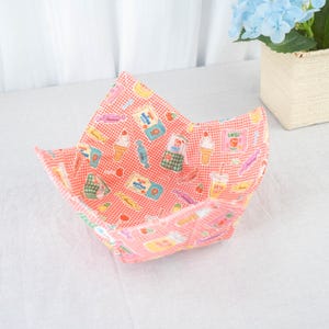 Op de afbeelding: Een vierkante stoffen kom met een rood-wit geblokt patroon. De kom is gevoerd met een kleurrijke print met ijsjes, snoepjes en melkpakken. De kom is ontworpen om voedsel in te bewaren.