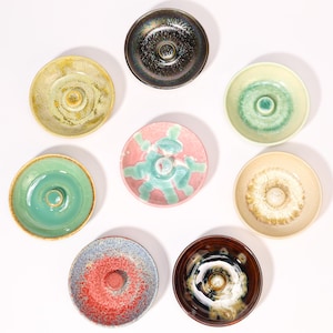 May include: A collection of eight ceramic incense holders in various colors and designs. The holders are round with a central raised post for holding incense sticks. Colors include green, pink, brown, and blue, with speckled and glazed finishes.