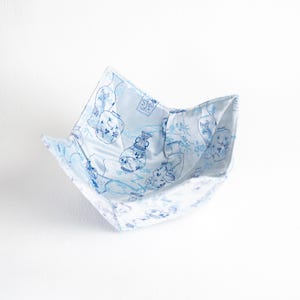 Könnte beinhalten: Eine hellblaue Stoffschale mit weißem Innenraum. Die Schale weist ein sich wiederholendes Muster aus blauen Vasen und floralen Designs auf. Die Schale ist offen und scheint aus einem weichen, faltbaren Material zu bestehen.